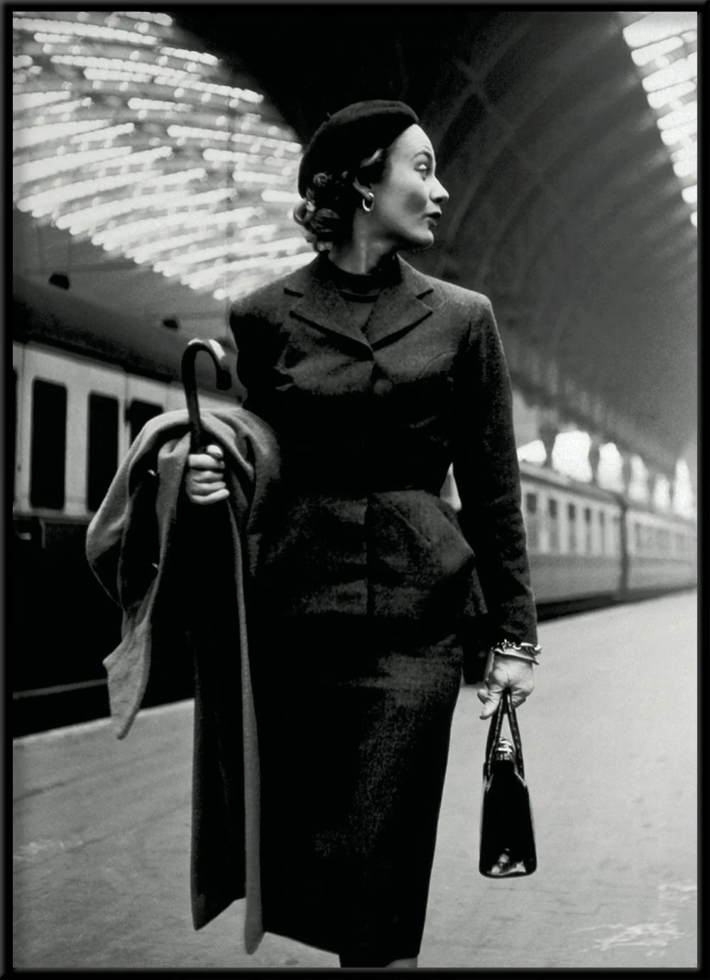 Black and white fashion photograph of a woman in a dark tailored coat and beret walking on a train station platform, holding a folded coat and a small handbag.