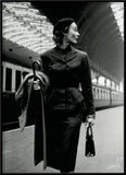 Black and white fashion photograph of a woman in a dark tailored coat and beret walking on a train station platform, holding a folded coat and a small handbag.