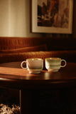 Two ceramic cups on a wooden table with a warm, dimly lit background.