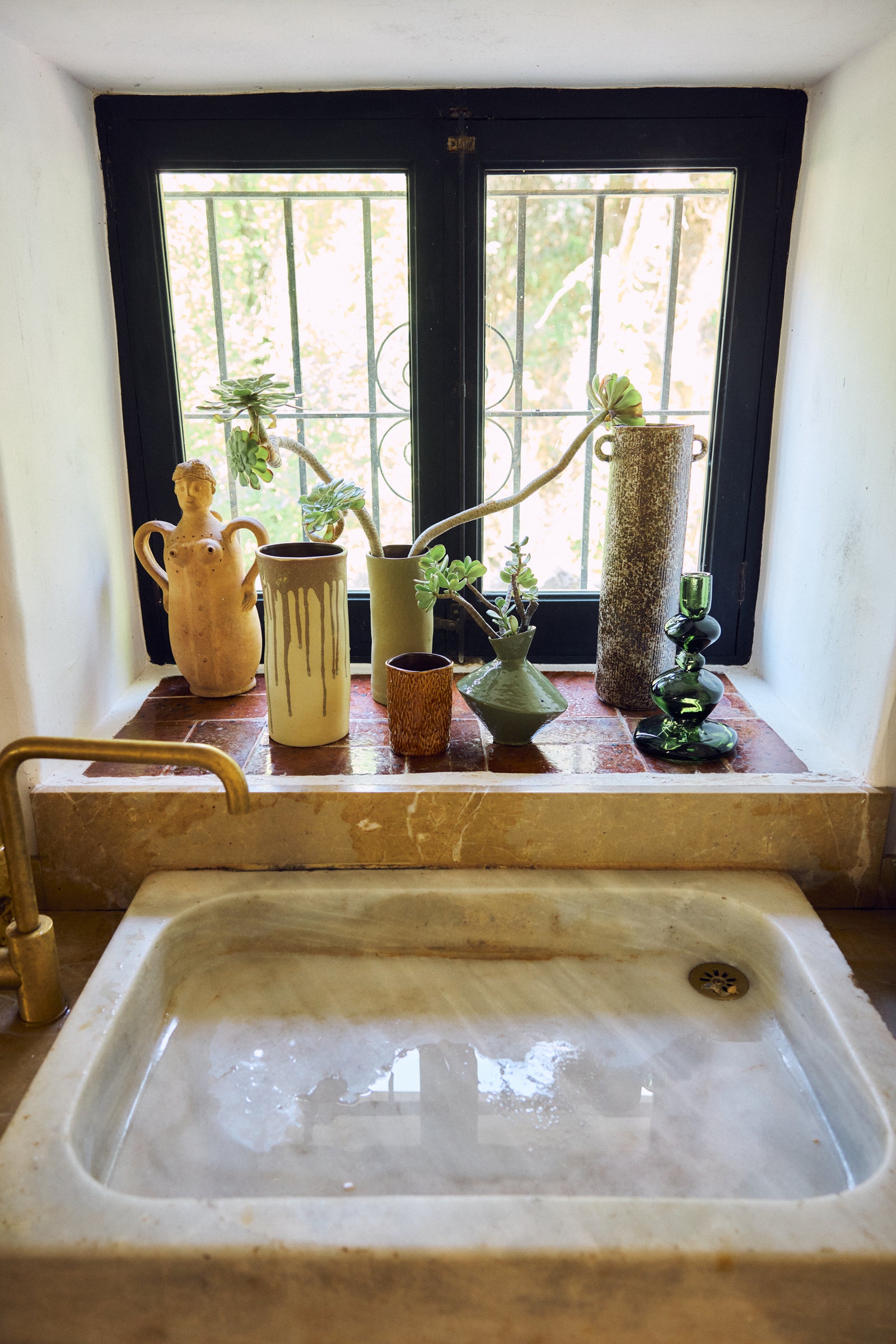 A rustic bathroom scene featuring a large white marble sink in the foreground and a matching stone window ledge with terracotta tiles, holding several vintage ceramic vases and plants. A brass faucet is visible on the left, and a black-framed window with metal bars is in the background.