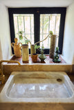 A rustic bathroom scene featuring a large white marble sink in the foreground and a matching stone window ledge with terracotta tiles, holding several vintage ceramic vases and plants. A brass faucet is visible on the left, and a black-framed window with metal bars is in the background.