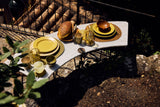 A styled outdoor dining table setup with yellow ceramic plates, matching yellow cups, textured bowls, and bread on a curved white stone bench, photographed in natural sunlight with surrounding foliage and rocks.