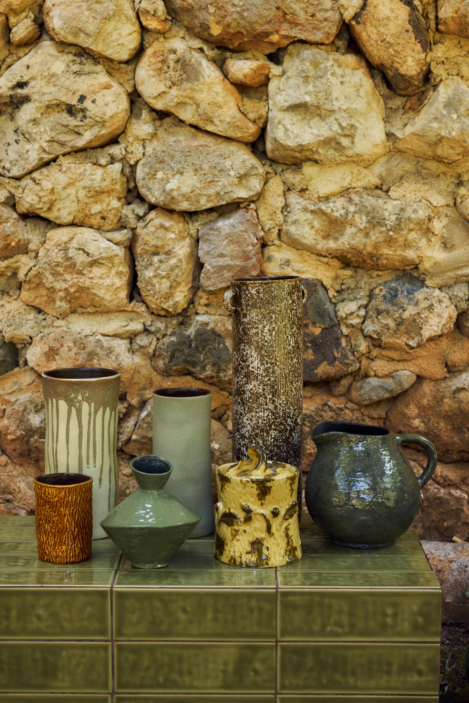 A collection of handmade ceramic vases and a pitcher displayed on a green tiled ledge in front of a rustic stone wall.