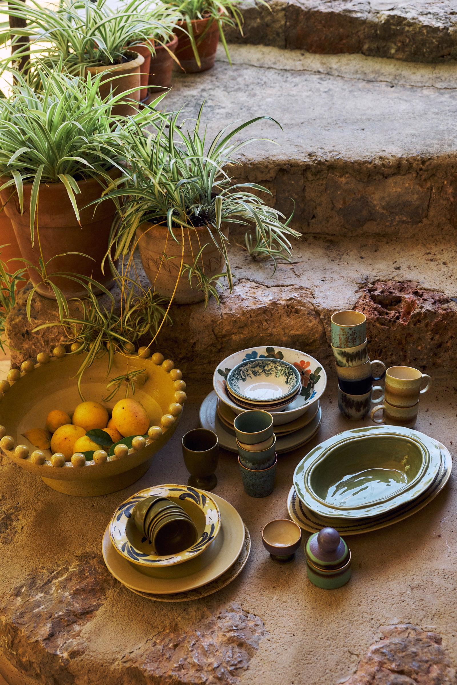 A collection of handmade ceramic tableware, including plates, bowls, cups, and stacked dish sets, arranged on rustic stone steps next to potted green plants and a bowl of oranges.