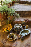 A collection of handmade ceramic tableware, including plates, bowls, cups, and stacked dish sets, arranged on rustic stone steps next to potted green plants and a bowl of oranges.