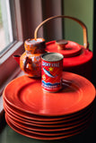 A stack of glossy red plates with a can of Lucky Star Picklards in Tomato Sauce in the center, set on a dark table near a window with a red teapot and ceramic pitcher in the background.