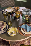 A styled dining table setting featuring a collection of hand-painted ceramic tableware, including a yellow floral bowl, patterned plates, green and amber glassware, and woven placemats on a dark olive tabletop.