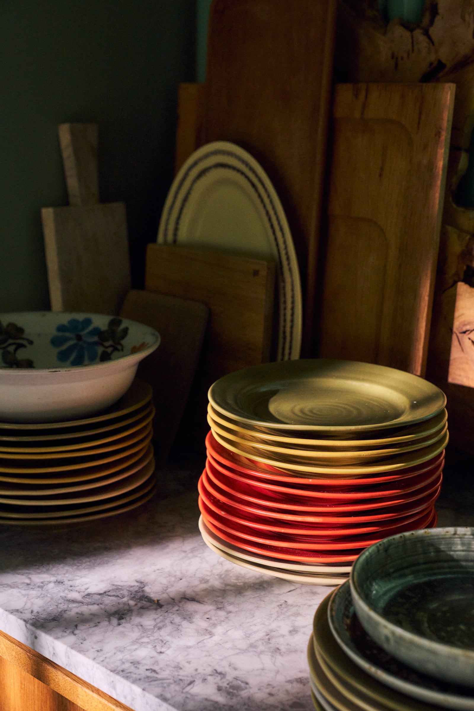 Stacks of ceramic plates in warm colors, including red, gold, and green, arranged on a marble countertop with wooden cutting boards and bowls in the background.