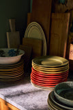 Stacks of ceramic plates in warm colors, including red, gold, and green, arranged on a marble countertop with wooden cutting boards and bowls in the background.