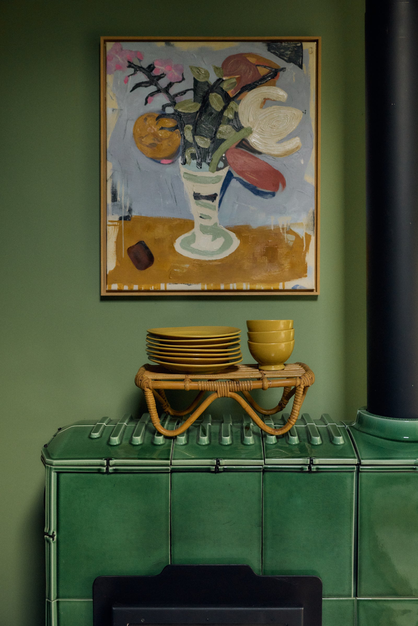 A framed abstract painting of a floral still life hangs on a dark green wall above a green tiled stove, with yellow plates and bowls on a small woven table.