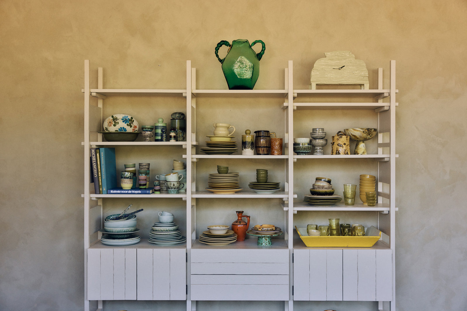A tall white wooden shelving unit with nine shelves, filled with various ceramic dishes, mugs, and decorative pottery, set against a textured beige plaster wall.