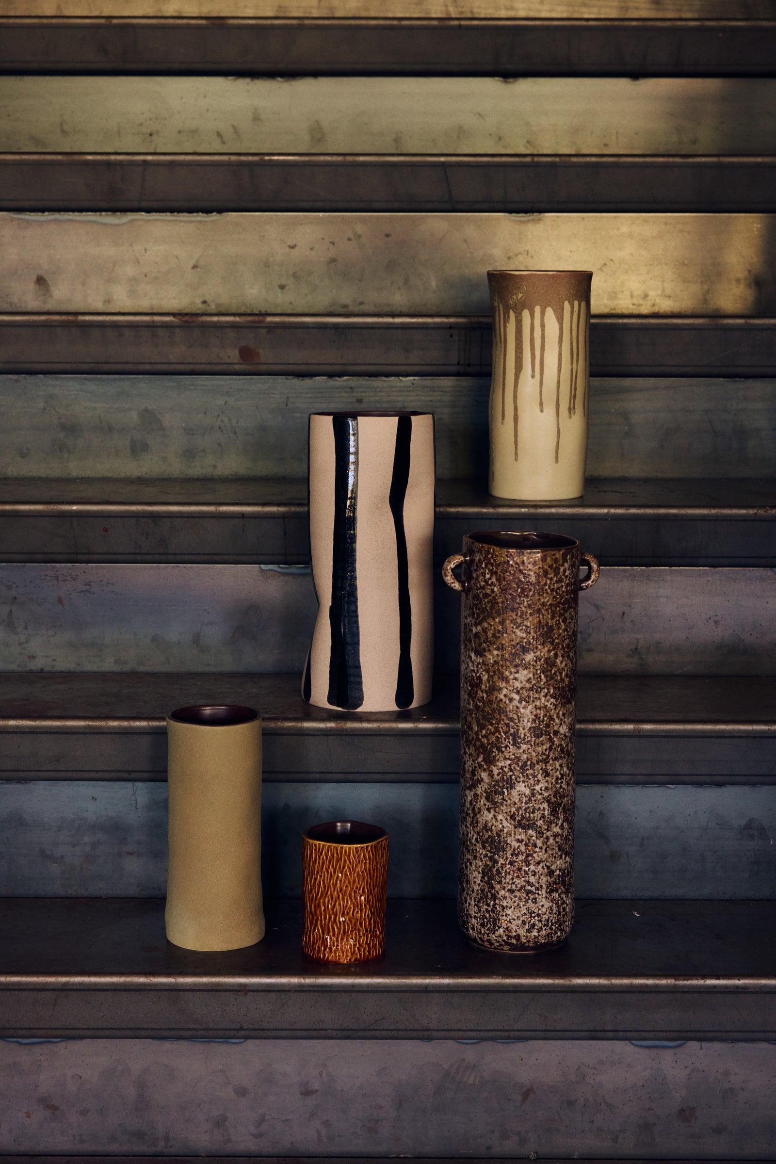 A collection of five handmade ceramic vases in various shapes and glazes, displayed on rustic wooden steps.