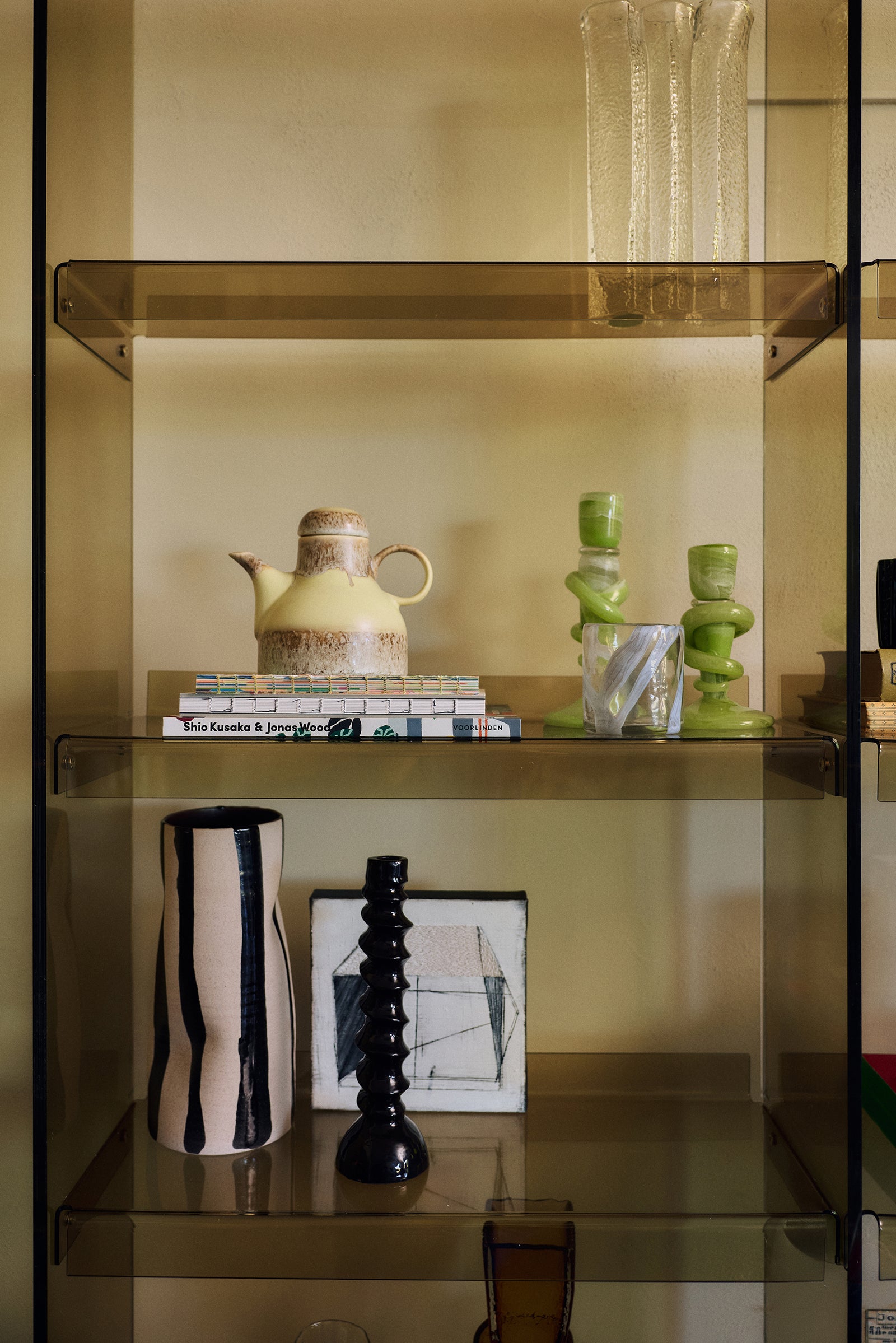 A modern glass shelving unit with a warm amber-brown tinted frame, styled with various decorative objects including ceramic vases, books, and a small teapot against a beige wall.