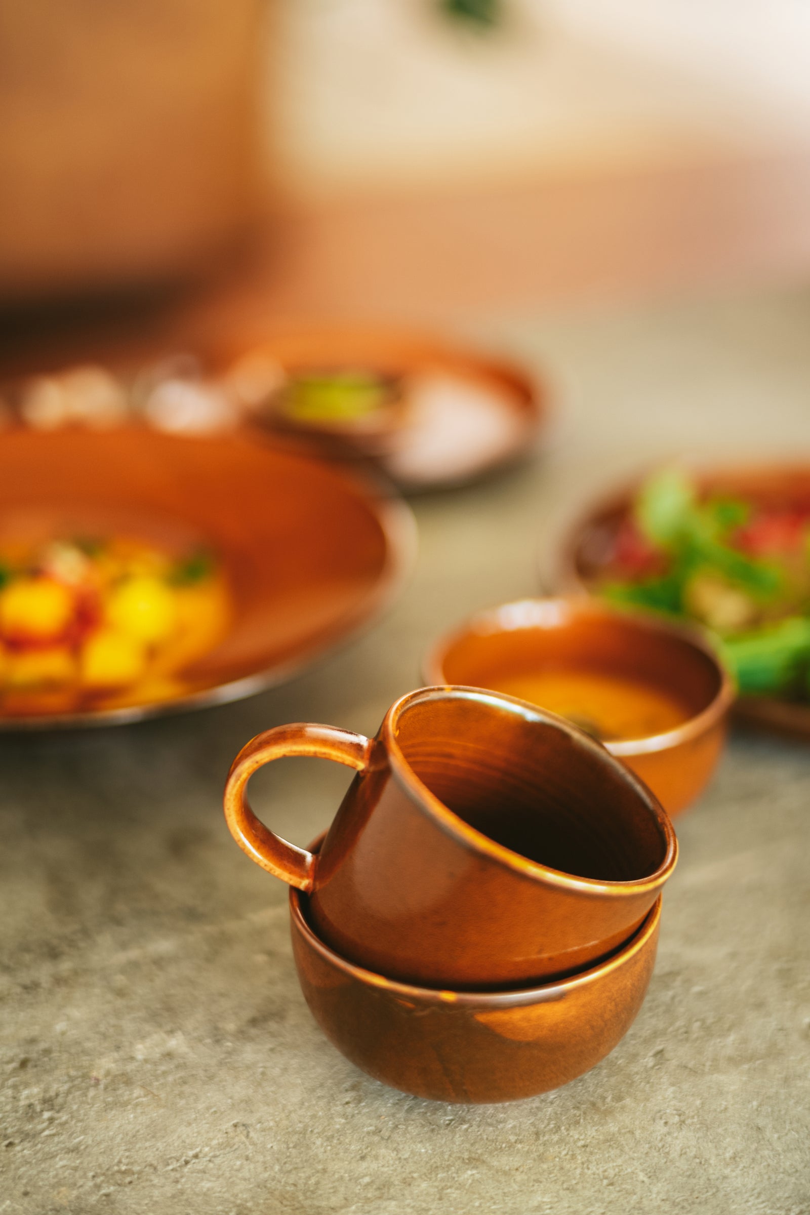 Stack of brown ceramic cups on a stone surface with blurred food items in the background