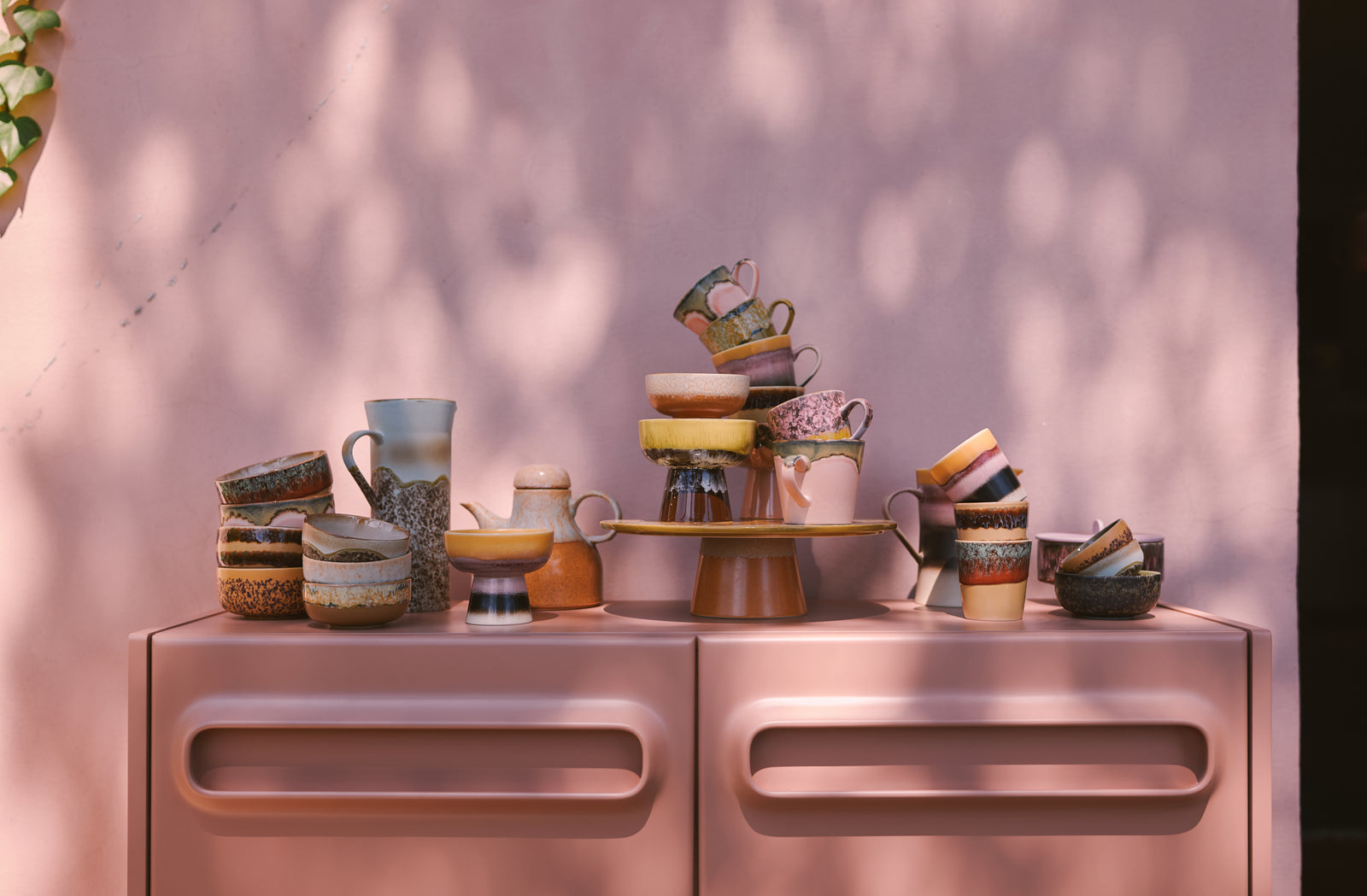 Collection of ceramic teapots and cups on a metallic surface with a pinkish-purple background