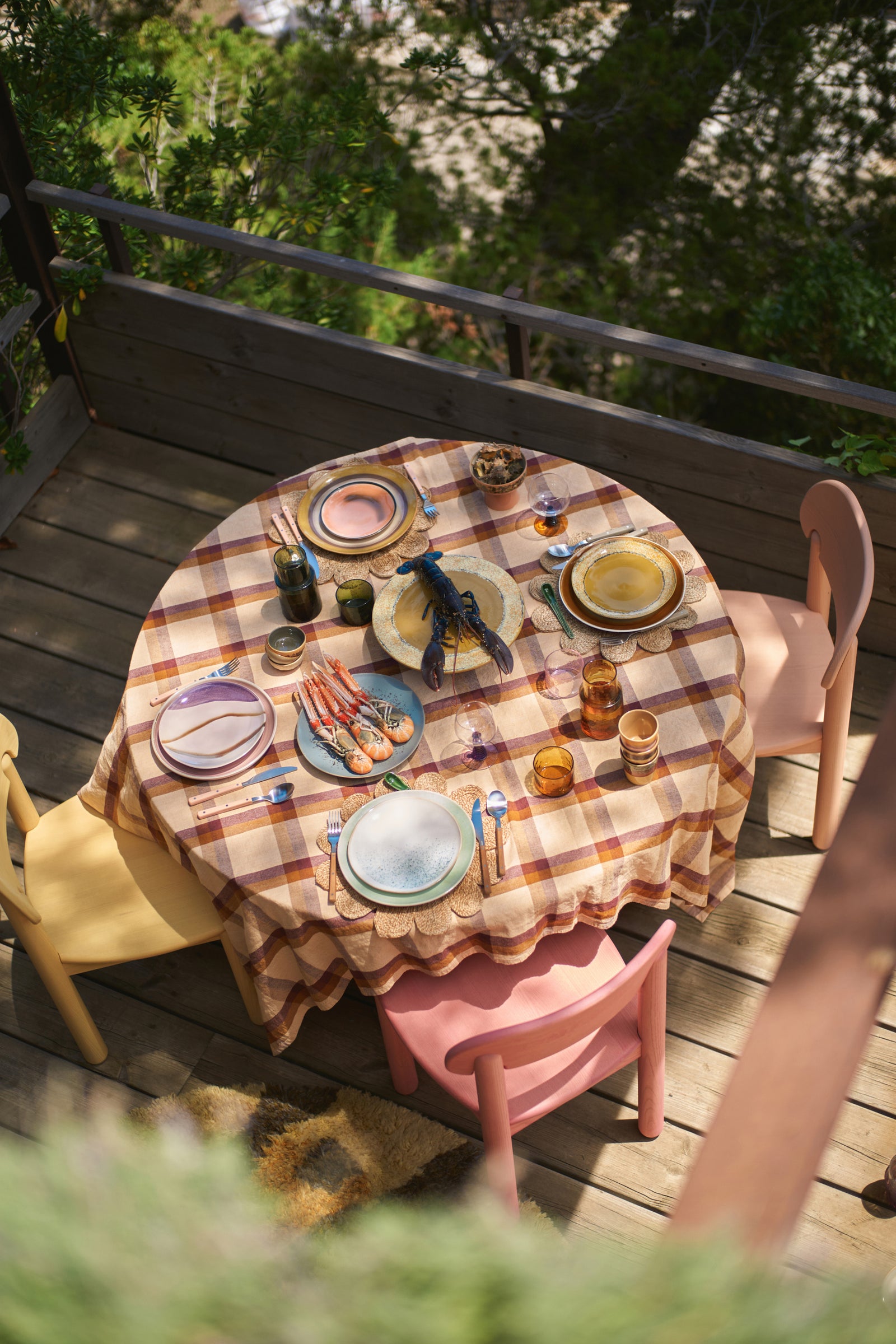 Dining table set with plates, cutlery, and glasses on a wooden deck with greenery in the background