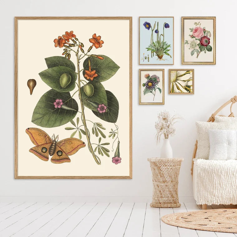 A large framed botanical art print featuring green leaves, orange flowers, and a butterfly, styled in a bright bedroom next to a white bed and smaller matching framed prints on the wall.
