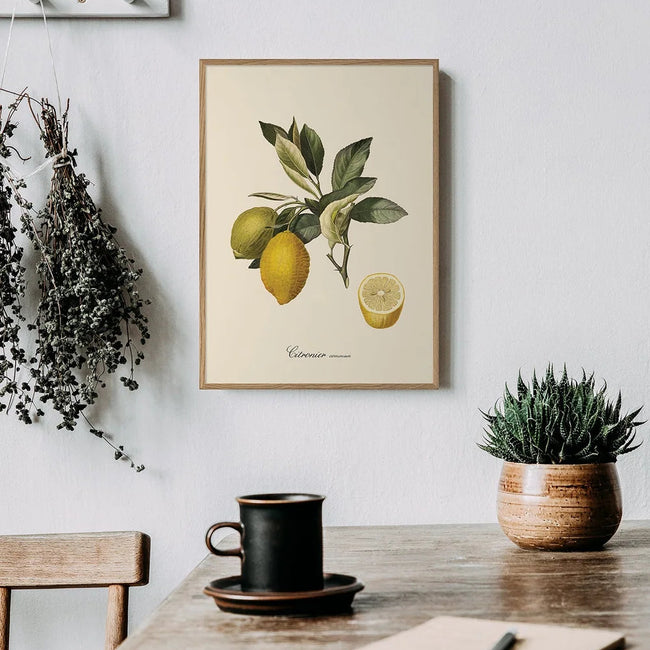 A framed botanical art print of lemons hanging on a light gray wall above a rustic wooden table with a black coffee cup, notebook, and a small potted succulent.