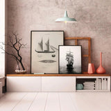 A styled interior scene with two framed black-and-white nautical ship prints displayed on a white bench against a textured gray wall. The room includes a hanging pendant light, a window on the left, decorative branches in a glass vase, books, and two tall coral-colored vases.