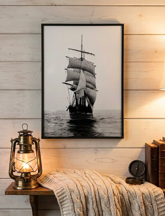 A framed black and white art print of a sailing ship on the ocean, displayed on a light wood paneled wall above a cozy shelf with a lit lantern, a cable-knit blanket, and a small clock.