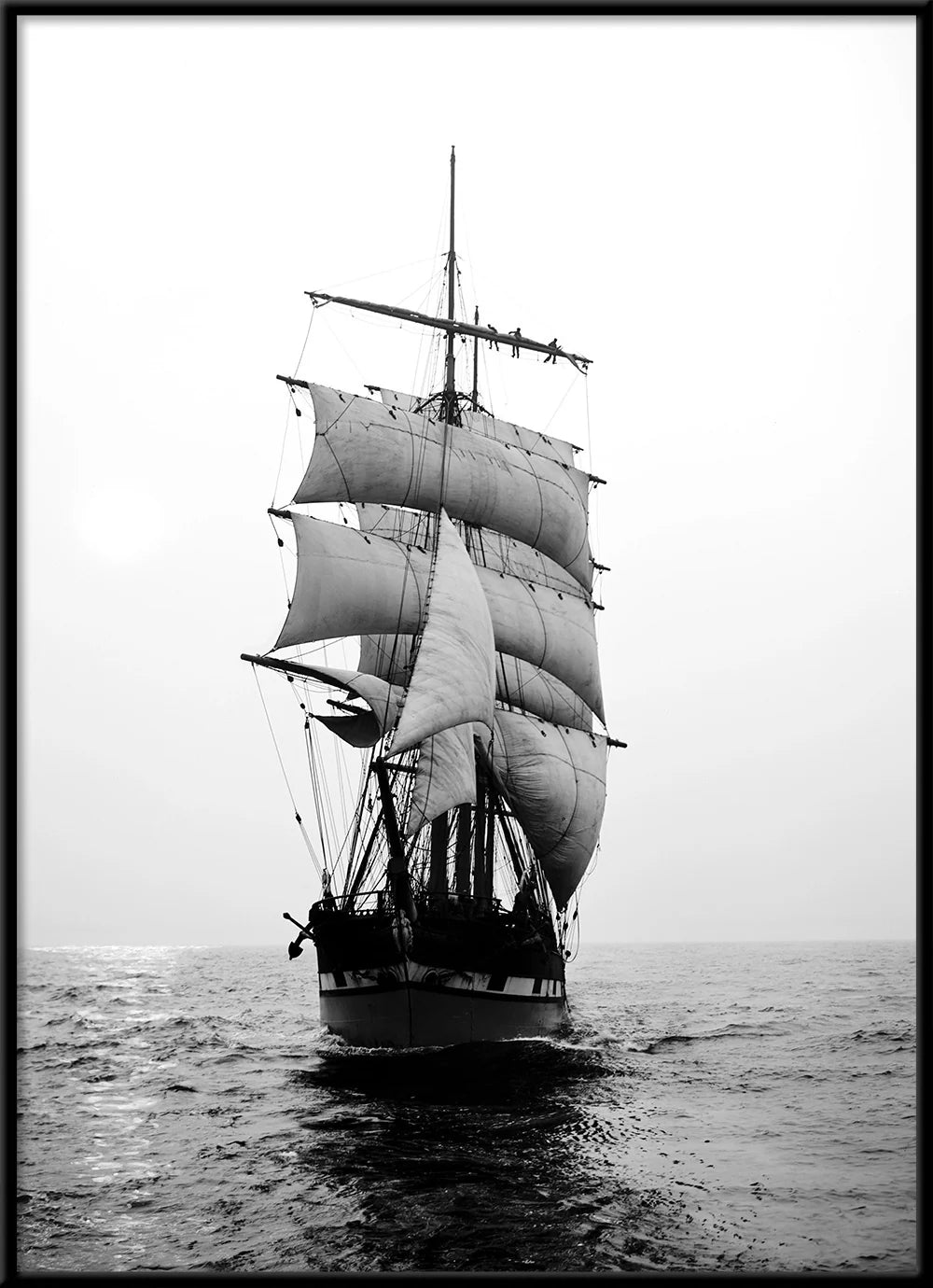 Black and white photograph of a tall-sailed historical sailing ship with multiple masts and billowing sails, sailing on open water under a cloudy sky.