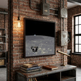 A framed space-themed artwork showing a lunar lander on the surface of the Moon with Earth visible in the black outer space background, enclosed in a light wood frame.