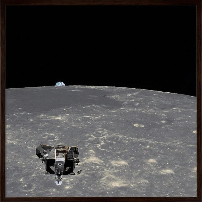 A framed space-themed artwork showing a lunar lander on the surface of the Moon with Earth visible in the black outer space background, enclosed in a light wood frame.