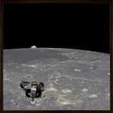 A framed space-themed artwork showing a lunar lander on the surface of the Moon with Earth visible in the black outer space background, enclosed in a light wood frame.