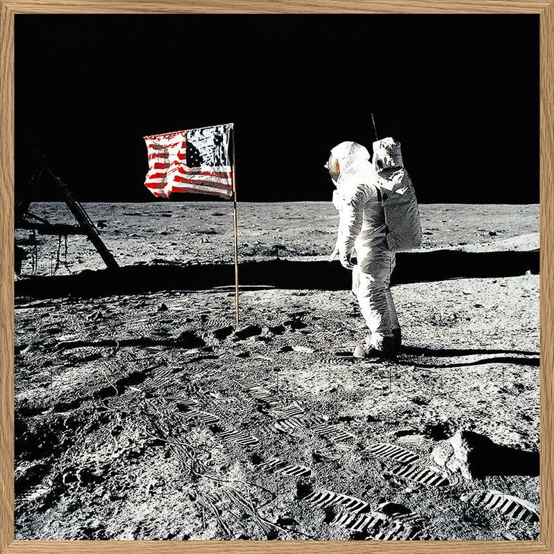 A framed square art print depicting a black-and-white astronaut standing on a dusty lunar surface next to an American flag, with a light wood frame border.