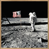A framed square art print depicting a black-and-white astronaut standing on a dusty lunar surface next to an American flag, with a light wood frame border.