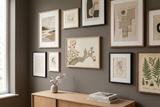 A gallery wall display of framed abstract and botanical art prints in neutral tones, mounted on a dark gray wall above a light wood console table with a vase and books.