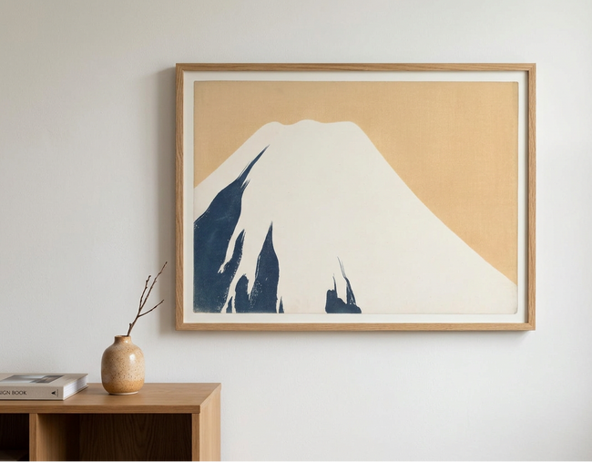 A framed minimalist art print of Mount Fuji, featuring a cream-colored mountain with dark blue shadows and a warm beige sky, enclosed in a light wood frame and hanging on a white wall above a wooden shelf.