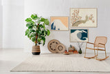 A styled interior scene with framed wall art above a low bench, including a large botanical print, a wave artwork, and smaller abstract pieces, alongside a potted plant, woven decor, books, a rattan chair, and a light rug in a bright room.
