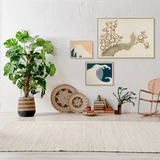 A styled interior scene with a large potted monstera plant on the left, framed wall art above, decorative plates and books, a small vase with greenery, and a wooden rocking chair on the right, all set against a white textured wall and light rug.