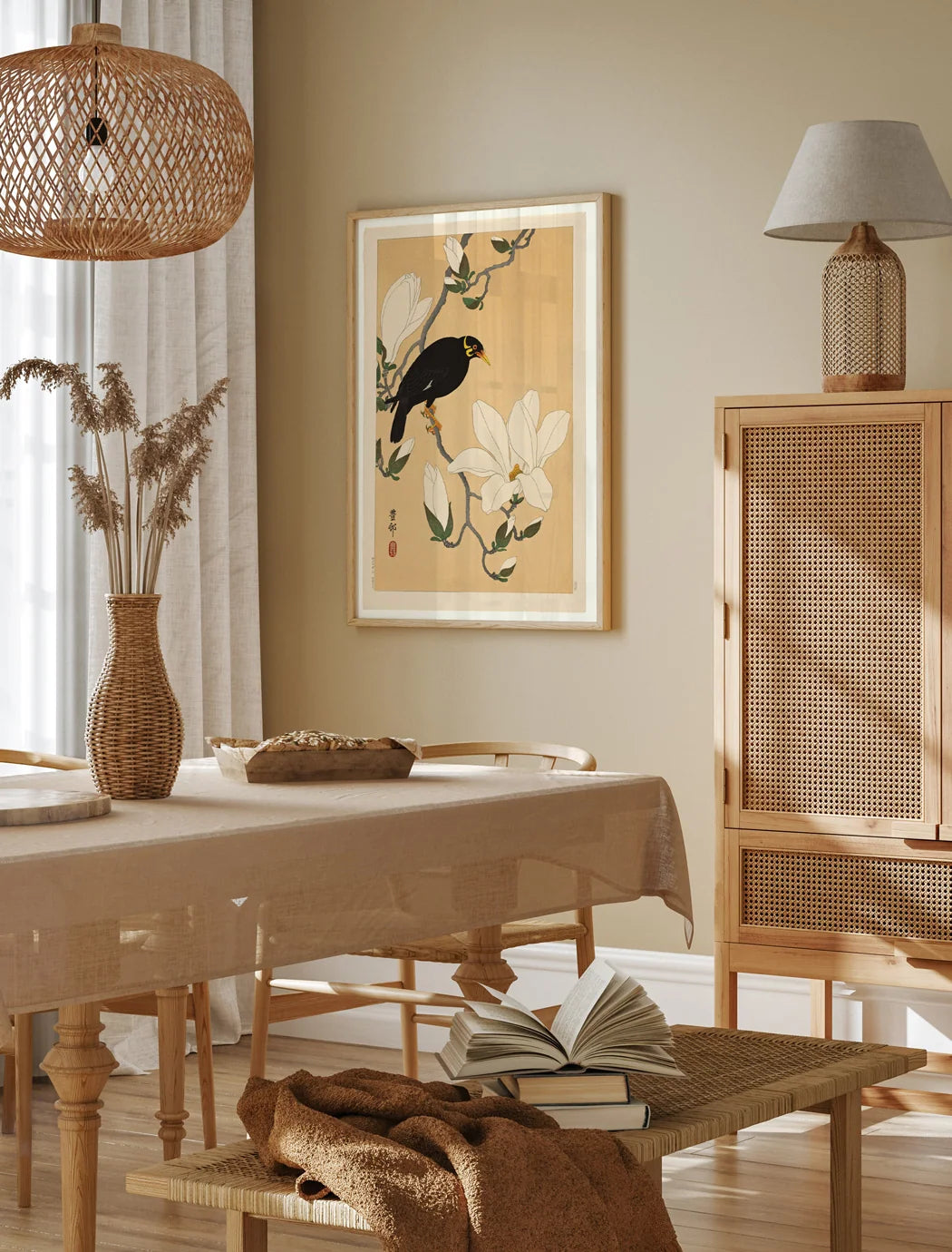 A framed art print featuring a black bird perched on white flowers against a warm beige background, displayed on a beige wall in a styled dining room setting with a wooden table, woven pendant light, and rattan cabinet.