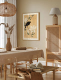 A framed art print featuring a black bird perched on white flowers against a warm beige background, displayed on a beige wall in a styled dining room setting with a wooden table, woven pendant light, and rattan cabinet.