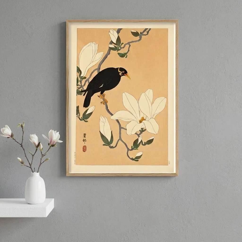 A framed art print featuring a black bird perched on a branch with white magnolia flowers, displayed on a light gray wall above a side table with a vase and a wooden chair.