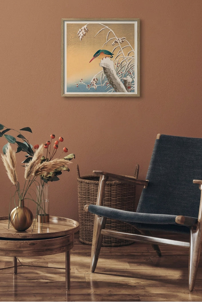 A framed Japanese-style art print featuring a green-and-blue bird perched on a snowy branch against a warm gold and blue background, displayed on a terracotta wall above a mid-century modern armchair and a small round side table with dried grasses.