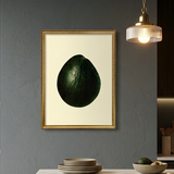 A framed art print featuring a dark green avocado on a cream background, hanging on a dark gray kitchen wall above a wooden island with marble countertop.