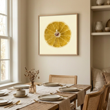 A framed wall art print of a yellow citrus slice on a cream background hangs above a rustic wooden dining table set with plates, chairs, and a table runner in a warm neutral dining room.