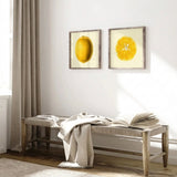Two framed lemon art prints hanging on a white wall above a wooden bench with a draped throw blanket and an open book in a bright room.