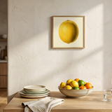 A framed botanical-style illustration of a single yellow lemon on a cream background, surrounded by a light wood frame.