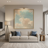 A large framed square wall art print depicting soft cream clouds against a pale blue sky, displayed above a light beige sofa in a modern, neutral-toned living room.
