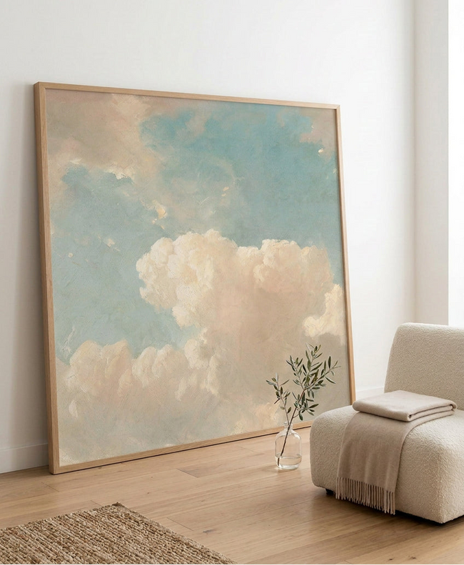 A framed art print depicting a soft, impressionistic sky with light blue and beige clouds, enclosed in a light wood frame.