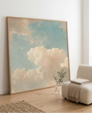A framed art print depicting a soft, impressionistic sky with light blue and beige clouds, enclosed in a light wood frame.