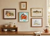 A styled interior wall with five framed art prints in different colors and styles, featuring a central vibrant orange-red crab print, flanked by fish, fruit, and lighthouse prints. Below the frames is a wooden shelf with bowls, a bottle, and a copper kettle on a light wood counter.