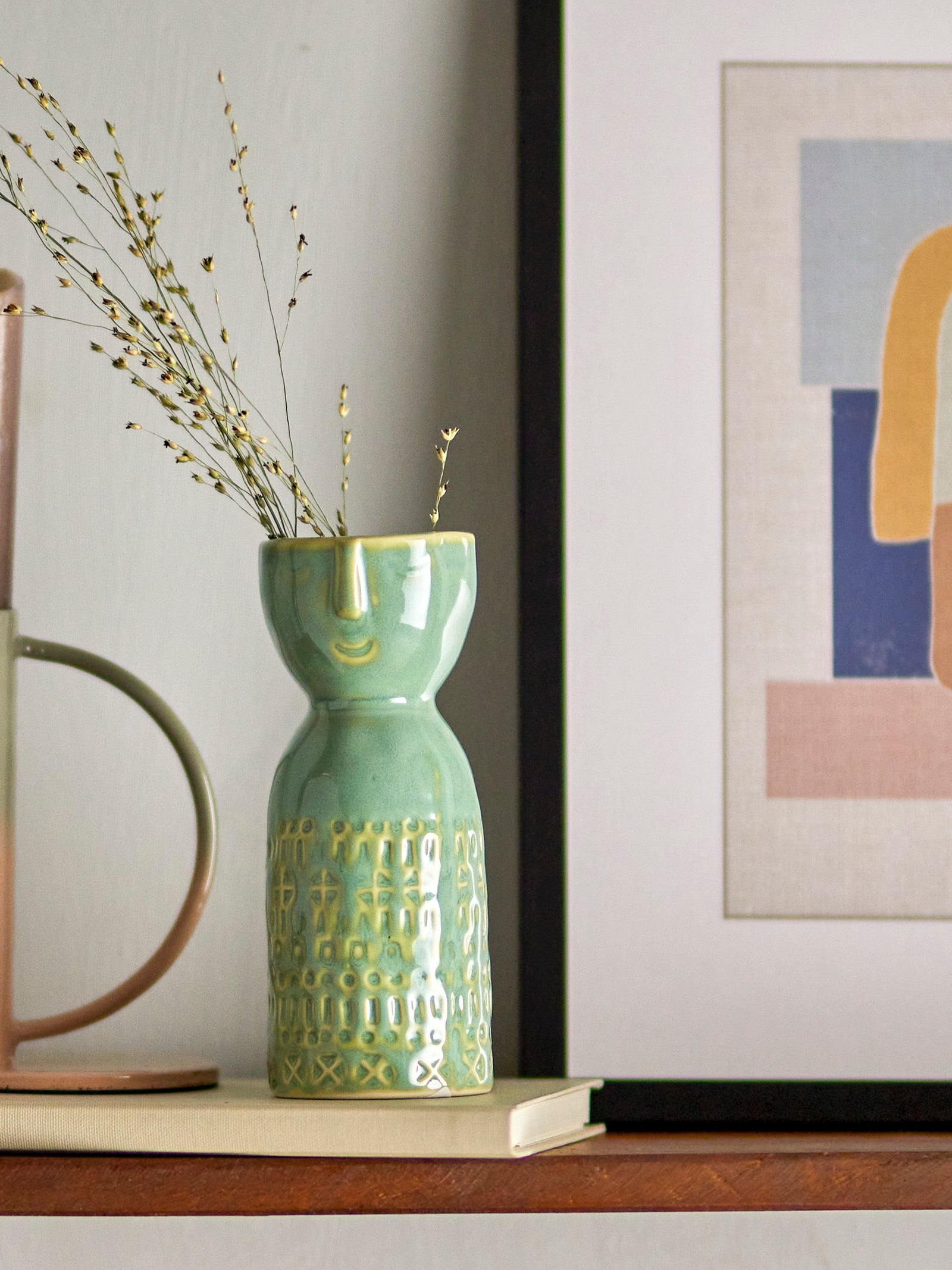 Green textured vase with dried plants on a shelf next to abstract art