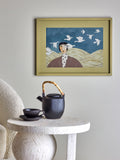 Framed artwork of a woman with birds on a blue sky background, placed above a table with a teapot and cup.