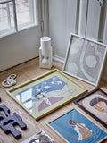 A styled interior scene showing several framed art prints and a textured white wall hanging leaning against a white door on a light wood floor. The most prominent item is a square framed print with a pale yellow-green frame, featuring an illustration of a woman with birds and a blue background.
