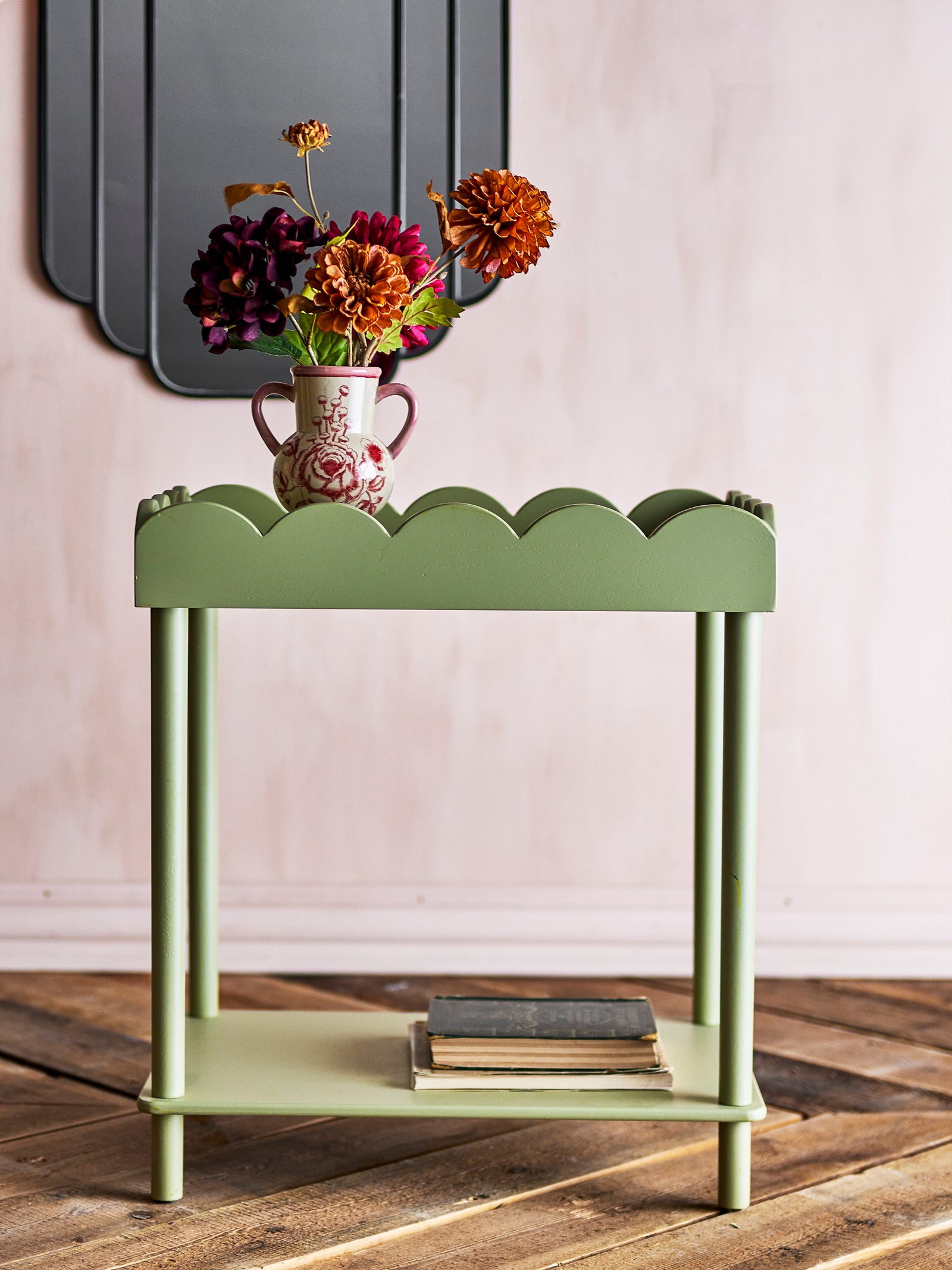 A sage green two-tier side table with a scalloped top edge, styled with a floral ceramic vase of dried flowers and a stack of books on a rustic wooden floor against a light textured wall.